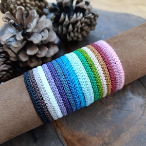 Puede incluir: Una colección de pulseras de crochet de colores en varios tonos de azul, verde, morado, marrón, blanco y rosa. Las pulseras están apiladas una encima de la otra, creando un efecto arcoíris.