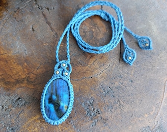 Puede incluir: Un collar de macramé azul con un colgante de labradorita ovalado de gran tamaño. El collar está hecho con un cordón trenzado y tiene un diseño sencillo.