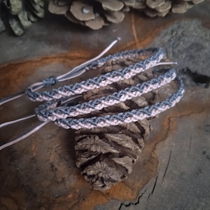 Peut inclure: Deux bracelets d'amitié en cordon gris et rose, tissés ensemble en un motif en croix. Les bracelets sont posés sur une surface en bois.