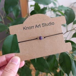 Op de afbeelding: Een dun zwart koordarmbandje met een enkele ronde paarse amethistkraal geflankeerd door twee kleine goudkleurige metalen kraaltjes. Het armbandje wordt gepresenteerd op een bruine kartonnen kaart met de tekst Knorr Art Studio.
