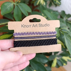 Op de afbeelding: Drie macramé armbanden in paars, zwart en een combinatie van paarse en zilveren kralen, gepresenteerd op een bruine kaart met de tekst "Knorr Art Studio". De armbanden worden voor een achtergrond van groene bladeren gehouden.
