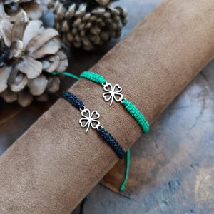 May include: Two friendship bracelets with a four-leaf clover charm. One bracelet is black and the other is green.