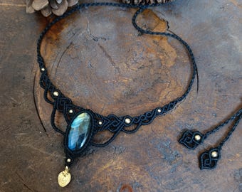 Puede incluir: Un collar gargantilla de macramé negro con un gran colgante de labradorita azul iridiscente y detalles dorados.