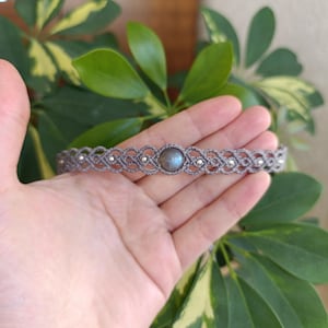 May include: A grey macramé choker necklace with a central, round, iridescent stone. The choker features a woven design with small silver-coloured beads. The necklace is displayed against a backdrop of green and yellow leaves.