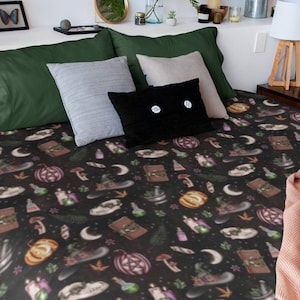 May include: A black bedspread with a repeating pattern of Halloween-themed illustrations, including pumpkins, witch hats, potion bottles, and moons. The bed is made up with green, gray, and black pillows, and a peach-colored blanket.