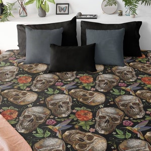 May include: A bed with a black skull-patterned comforter, surrounded by black and gray pillows. The comforter features detailed skull illustrations, flowers, and hummingbirds. The room is decorated with plants and a lamp.