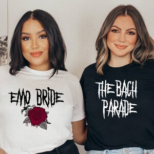 May include: Two women wearing graphic t-shirts. One shirt is white with the text "EMO BRIDE" and a red rose graphic. The other shirt is black with the text "THE BACH PARADE" in white. Both shirts are short-sleeved.