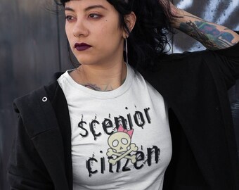 May include: White t-shirt with the text "$cenior Citizen" and a skull graphic with a pink bow. The person is wearing a black jacket and a plaid skirt. The background is a dark wall with graffiti.