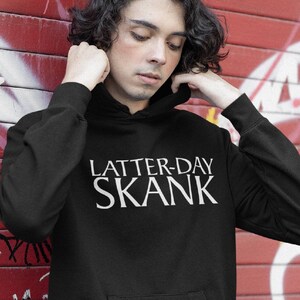 May include: Black hoodie with the text "LATTER-DAY SKANK" in white. The hoodie has a front pocket and a drawstring hood. The person in the image is wearing the hoodie and is pulling up the hood.