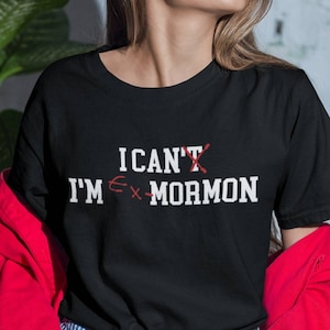 May include: Black t-shirt with the text "I can't, I'm ex-Mormon" in white. The word "can't" has a red X through it, and the "M" in "Mormon" is crossed out. The person is wearing a red jacket and blue and white striped jeans.