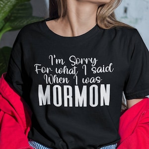 May include: Black t-shirt with the text "I'm Sorry For what I said When I was MORMON" in white. The shirt is worn with blue and white striped jeans and a red jacket.