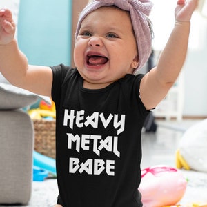 May include: A black baby bodysuit with the text "HEAVY METAL BABE" in white. The baby is kneeling on the floor with arms raised and mouth open. The baby is wearing a light purple headband.