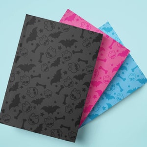 May include: Three notebooks with patterned covers. The first notebook is black with a repeating pattern of skulls, bats, bones, and stars. The second notebook is pink, and the third is blue. All three notebooks have the same pattern.