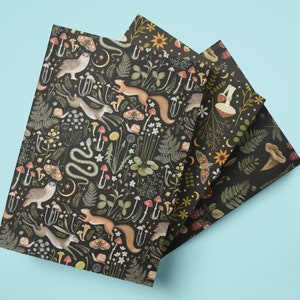 May include: Three notebooks with a dark background and a whimsical forest pattern. The pattern includes owls, rabbits, squirrels, mushrooms, and other forest elements. The notebooks are fanned out on a light blue surface.