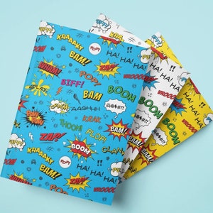 May include: Three notebooks with comic book-style covers. The covers feature action words like "BAM!" and "ZAP!" in speech bubbles. The notebooks are blue, white, and yellow, with a light blue background.