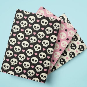 May include: Three notebooks with skull patterns in black, pink, and cream colors. The black notebook has white skulls and pink hearts. The pink notebook has white skulls and black stars. The cream notebook has black skulls.