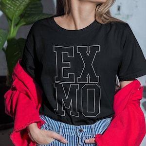 May include: Black t-shirt with the text "EX MO" in a white, collegiate-style font. The person is wearing blue and white striped jeans and a red jacket. The background includes a green plant and a white brick wall.