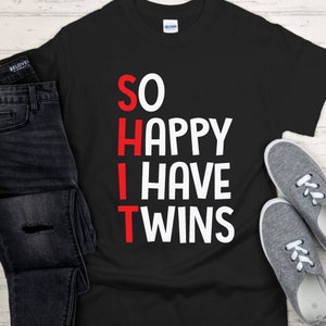 May include: Black t-shirt with the text "SO HAPPY I HAVE TWINS" in white and red lettering. The shirt is displayed with a pair of gray sneakers and a pair of dark blue jeans.