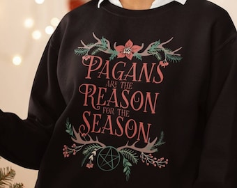 May include: Black sweatshirt with the text "Pagans are the Reason for the Season" in pink and green. The design includes floral and antler elements. The person is standing near a decorated Christmas tree.