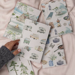 May include: A collection of notebooks with watercolor illustrations of camping and travel themes. Designs include camper vans, cabins, campfires, and outdoor gear. The notebooks are arranged on a soft, pink blanket.