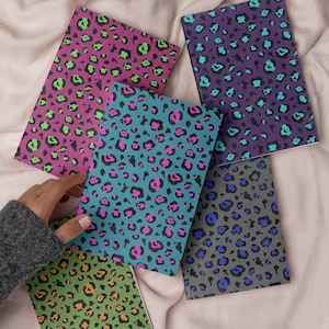 May include: A collection of notebooks with colorful leopard print designs. The notebooks feature various color combinations, including pink and green, teal and pink, purple and teal, gray and blue, and green and orange. The notebooks are arranged on a soft, cream-colored fabric.