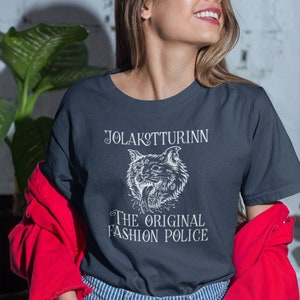 May include: Navy blue t-shirt with the text "JOLAKOTTURINN" above a white illustration of a cat's head, and the words "THE ORIGINAL FASHION POLICE" below. The person is wearing a red jacket and blue and white striped jeans.