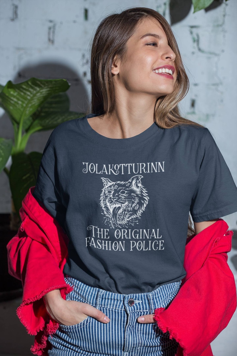 May include: Navy blue t-shirt with the text "JOLAKOTTURINN" above a white illustration of a cat's head, and the words "THE ORIGINAL FASHION POLICE" below. The person is wearing a red jacket and blue and white striped jeans.
