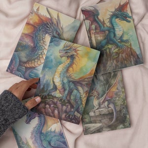 May include: A collection of five notebooks featuring watercolor illustrations of dragons in various poses. The dragons are depicted in shades of blue, green, and purple, with detailed wings and scales. The notebooks are on a soft, pinkish-beige surface.