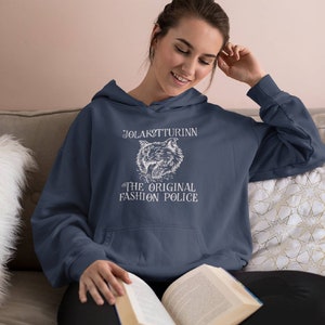 May include: Navy blue hoodie with a white graphic featuring a wolf and the text "JOLAKOTTURINN THE ORIGINAL FASHION POLICE." The hoodie has a drawstring hood and a front pocket. The person in the image is reading a book.