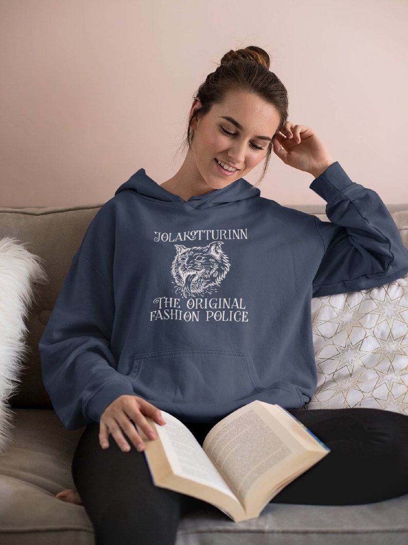 May include: Navy blue hoodie with a white graphic featuring a wolf and the text "JOLAKOTTURINN THE ORIGINAL FASHION POLICE." The hoodie has a drawstring hood and a front pocket. The person in the image is reading a book.