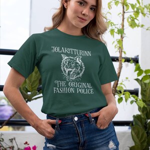 May include: Dark green t-shirt with white text and a wolf illustration. The text reads "JOLAKOTTURINN" above the wolf and "THE ORIGINAL FASHION POLICE" below. The model is wearing a denim skirt.