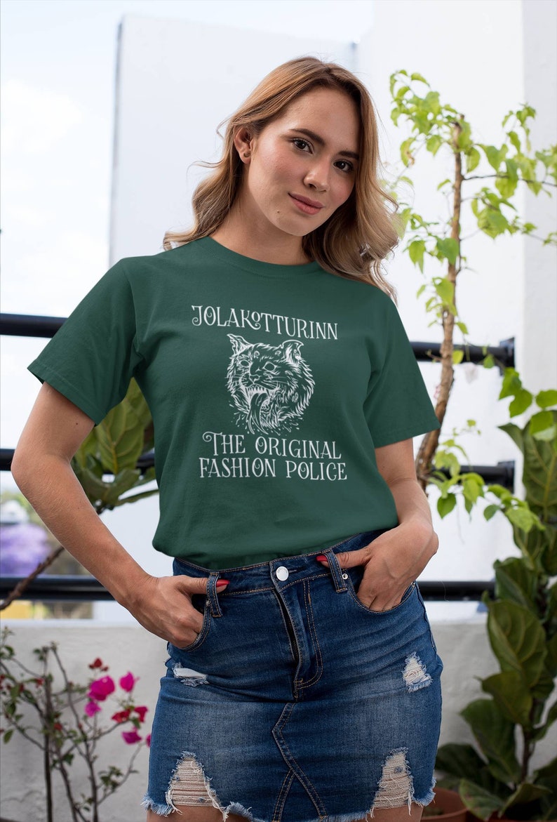 May include: Dark green t-shirt with white text and a wolf illustration. The text reads "JOLAKOTTURINN" above the wolf and "THE ORIGINAL FASHION POLICE" below. The model is wearing a denim skirt.