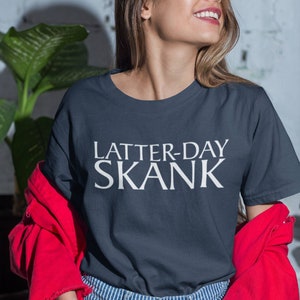 May include: A navy blue t-shirt with the text "LATTER-DAY SKANK" in white block letters. The person is wearing a red jacket and blue and white striped jeans. The background includes a green plant and a white brick wall.