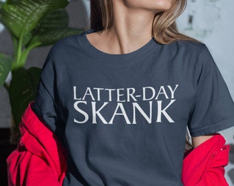 May include: A navy blue t-shirt with the text "LATTER-DAY SKANK" in white block letters. The person is wearing a red jacket and blue and white striped jeans. The background includes a green plant and a white brick wall.
