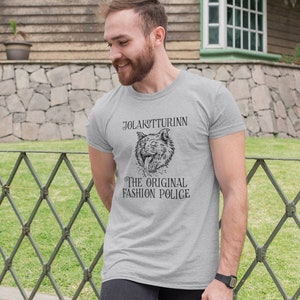 May include: A light gray t-shirt featuring a detailed black illustration of a wolf's head with the text "JOLAKOTTURINN THE ORIGINAL FASHION POLICE". The shirt is made of a soft material.