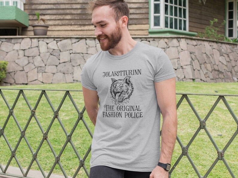 May include: A light gray t-shirt featuring a detailed black illustration of a wolf's head with the text "JOLAKOTTURINN THE ORIGINAL FASHION POLICE". The shirt is made of a soft material.