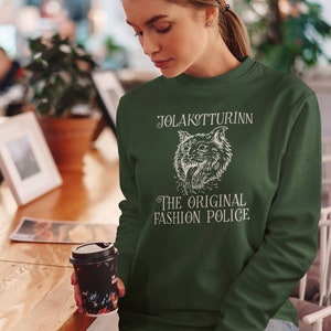 May include: Dark green sweatshirt with white text and a graphic of a snarling cat. The text reads "JOLAKOTTURINN THE ORIGINAL FASHION POLICE". The sweatshirt is long-sleeved and has a crew neck.