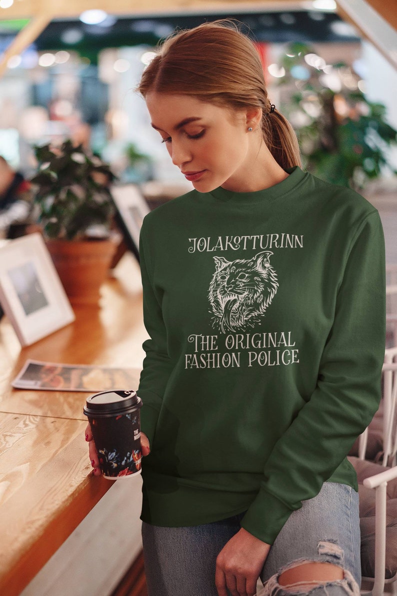 May include: Dark green sweatshirt with white text and a graphic of a snarling cat. The text reads "JOLAKOTTURINN THE ORIGINAL FASHION POLICE". The sweatshirt is long-sleeved and has a crew neck.