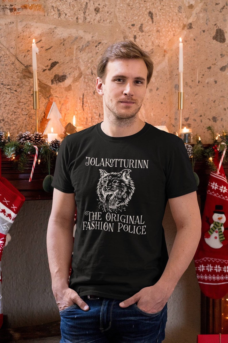 May include: Black t-shirt with the text "JOLAKOTTURINN THE ORIGINAL FASHION POLICE" and a detailed cat illustration. The person is wearing blue jeans. The background includes a fireplace with candles and Christmas decorations.