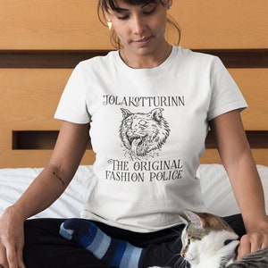 May include: White t-shirt with a black graphic of a snarling cat and the text "JOLAKOTTURINN THE ORIGINAL FASHION POLICE". The shirt is made of a soft material.