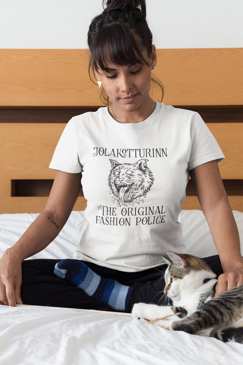 May include: White t-shirt with a black graphic of a snarling cat and the text "JOLAKOTTURINN THE ORIGINAL FASHION POLICE". The shirt is made of a soft material.