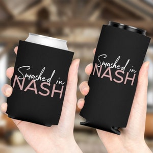 May include: Two black can coolers with the text "Smashed in NASH" in white and pink script. One cooler holds a silver can, the other is empty. The coolers are held in hands.