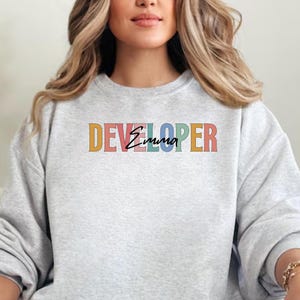 Puede incluir: Una sudadera gris claro con la palabra "DEVELOPER" en letras de colores y el nombre "Emma" en cursiva. La sudadera es de cuello redondo.