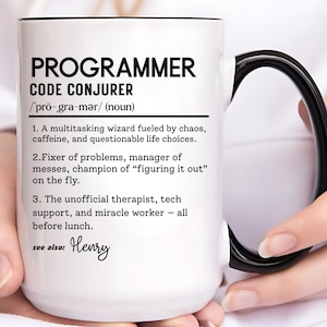 Puede incluir: Taza de cerámica blanca con asa y borde negros. La taza presenta el texto "PROGRAMMER CODE CONJURER" con una definición. La taza está sujeta por una persona.