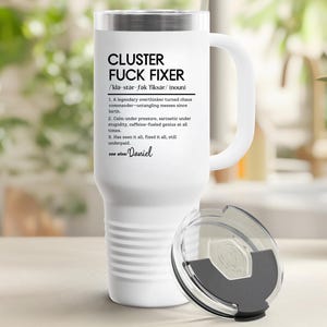 Op de afbeelding: Witte reismok met een handvat en een zilveren deksel. De mok heeft de tekst "CLUSTER FUCK FIXER" met een definitie. De mok is ontworpen om dranken warm of koud te houden.