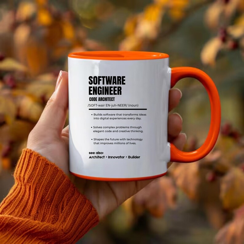 K&ouml;nnte beinhalten: Ein wei&szlig;er Keramikbecher mit leuchtend orangefarbenem Innenraum und Henkel. Die Vorderseite zeigt schwarzen Text, der einen Softwareentwickler als Code-Architekten definiert, inklusive phonetischer Aussprache und Aufgabenbeschreibungen.