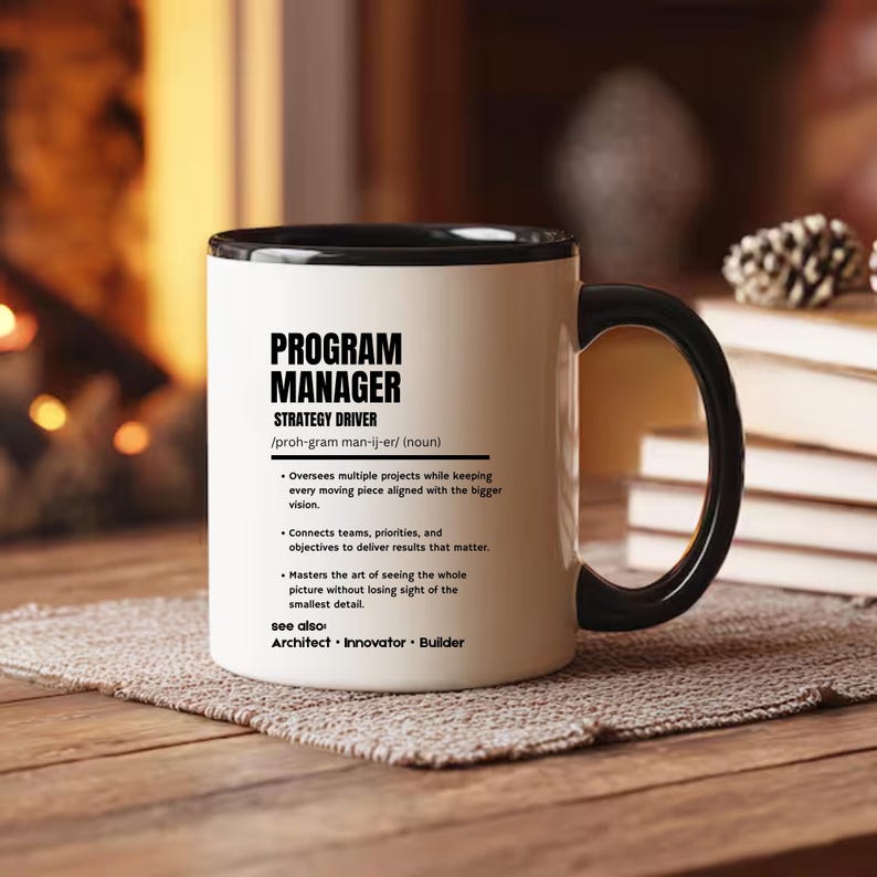 May include: A white ceramic mug with a black handle and interior rim. The front features black text defining a Program Manager as a strategy driver, including phonetic spelling, bulleted job responsibilities, and related terms like Architect, Innovator, and Builder.