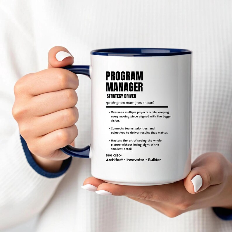 May include: A white ceramic mug with a dark blue handle and interior rim. The mug features black printed text defining a Program Manager as a strategy driver, including bulleted responsibilities and a see also section listing Architect, Innovator, and Builder.