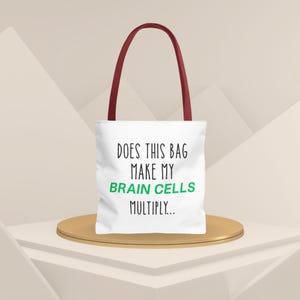May include: White tote bag with burgundy handles, featuring the text "DOES THIS BAG MAKE MY BRAIN CELLS MULTIPLY..." in black and green. The bag is displayed on a gold-colored platform.