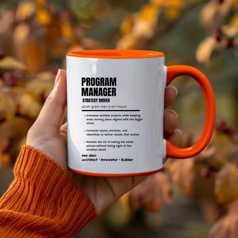 May include: A white ceramic mug with a bright orange interior and handle. The front features black text defining a Program Manager as a strategy driver, including phonetic pronunciation, bulleted job responsibilities, and related terms.